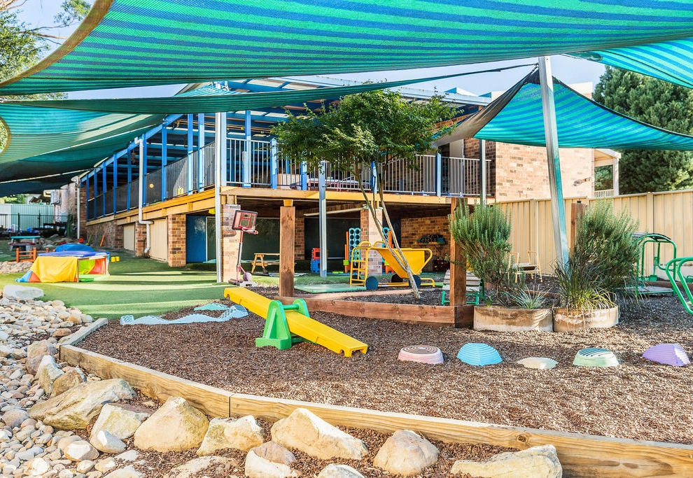 Outdoor playground with various equipment including a seesaw, climbing structure, and shaded areas. Surrounded by a garden and rocks, with a building and greenery in the background.