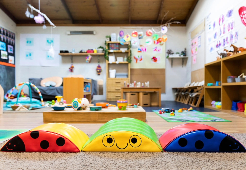 Colorful and playful preschool classroom featuring ladybug-themed soft mats, various educational toys, crafts on display, and a cozy reading area.