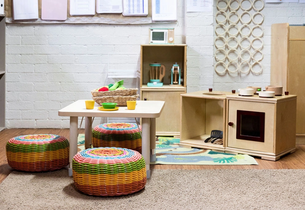 Children's playroom with a small white table, colorful woven stools, toy kitchen appliances, and a woven basket with toy vegetables on a patterned rug. Shelves and wall decorations are in the background.