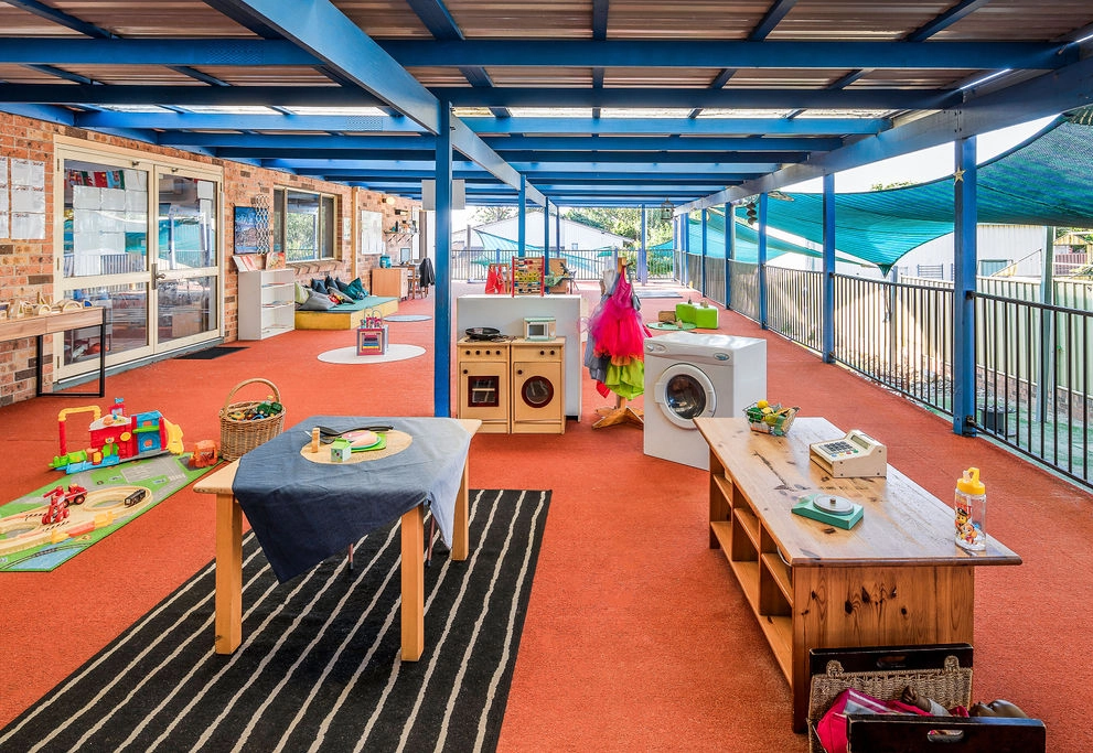 A covered outdoor play area features a table with a blue cloth, toy appliances, a play kitchen set, a washing machine, toy blocks, and activity stations.