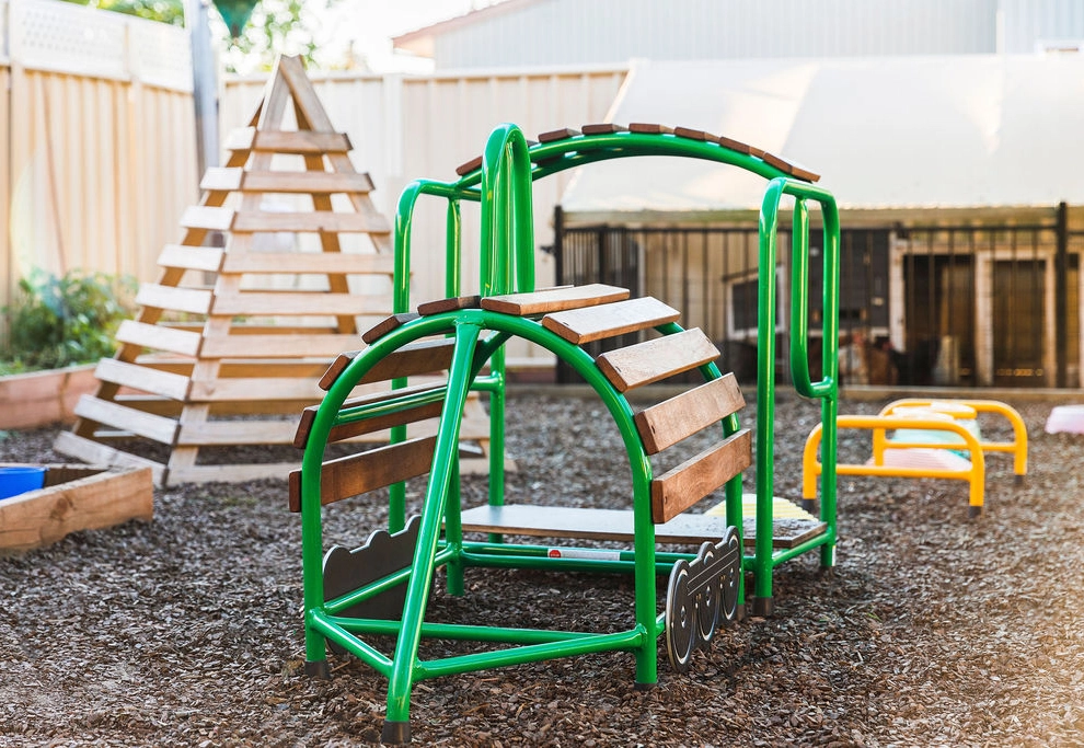 Outdoor playground featuring a green and brown play structure with a climbing arch and attached slide. A wooden pyramid structure stands in the background. The ground is covered in wood chips.