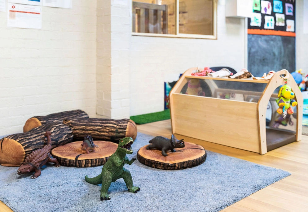 A children's play area features a blue rug with toy dinosaurs on wooden stumps and a wooden storage unit filled with stuffed animals in the background.