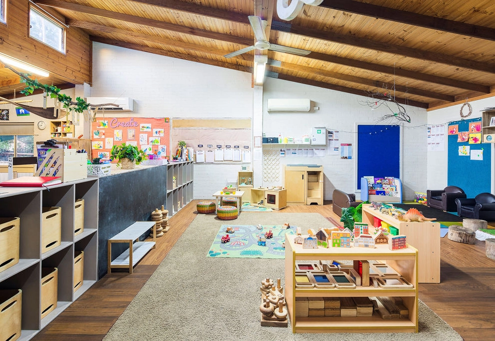 A well-lit, organized preschool classroom with wooden shelves, cubbies, toys, art supplies, and colorful decorations. A soft rug with play areas is in the center, and children's art adorns the walls.