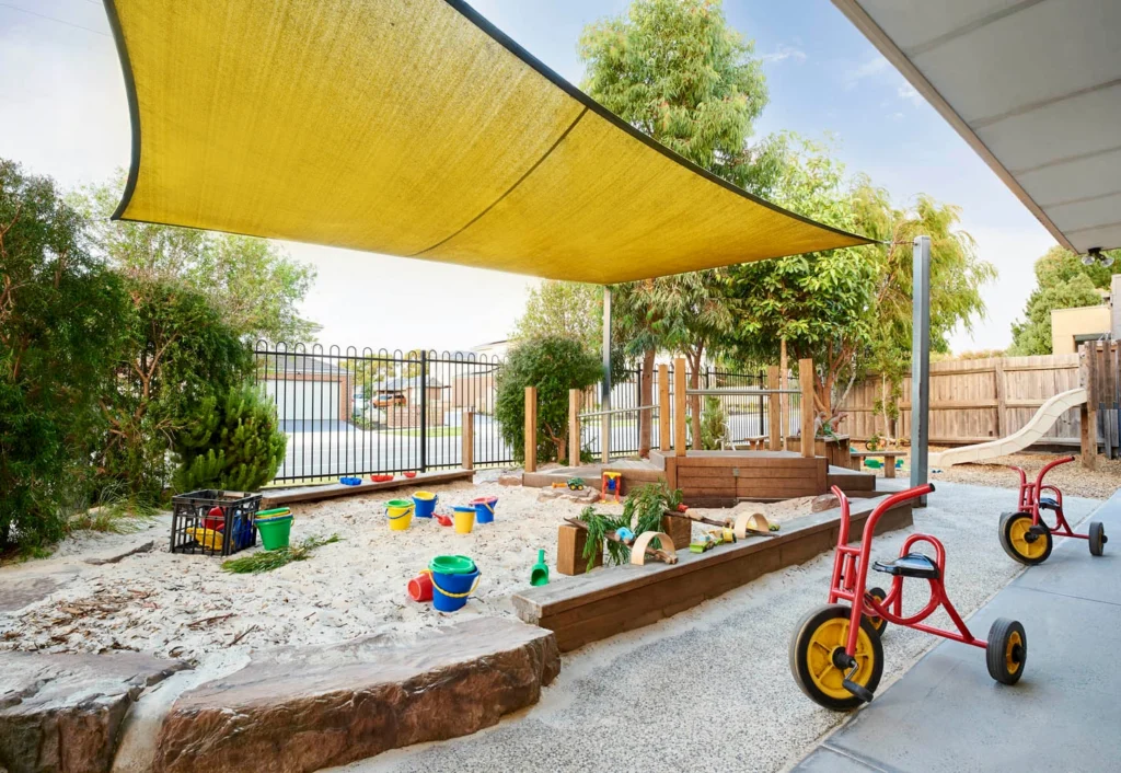 Outdoor children's play area with sandbox, plastic toys, tricycles, and a yellow shade canopy. Perfect for early learning at a kindergarten or childcare center, it’s enclosed by a black metal fence and surrounded by greenery and wooden structures.