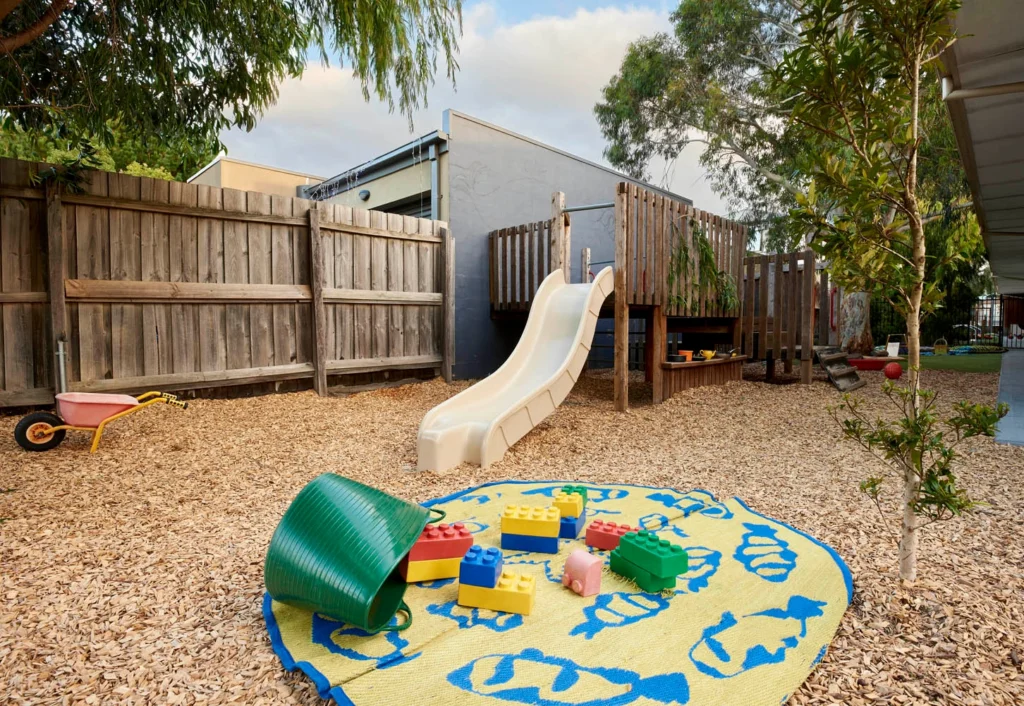 A playground at the day care features a white slide attached to a wooden play structure, a colorful play mat with large toy blocks, a green barrel, and a small wheelbarrow on a wood chip surface, creating an ideal setting for early learning and development.