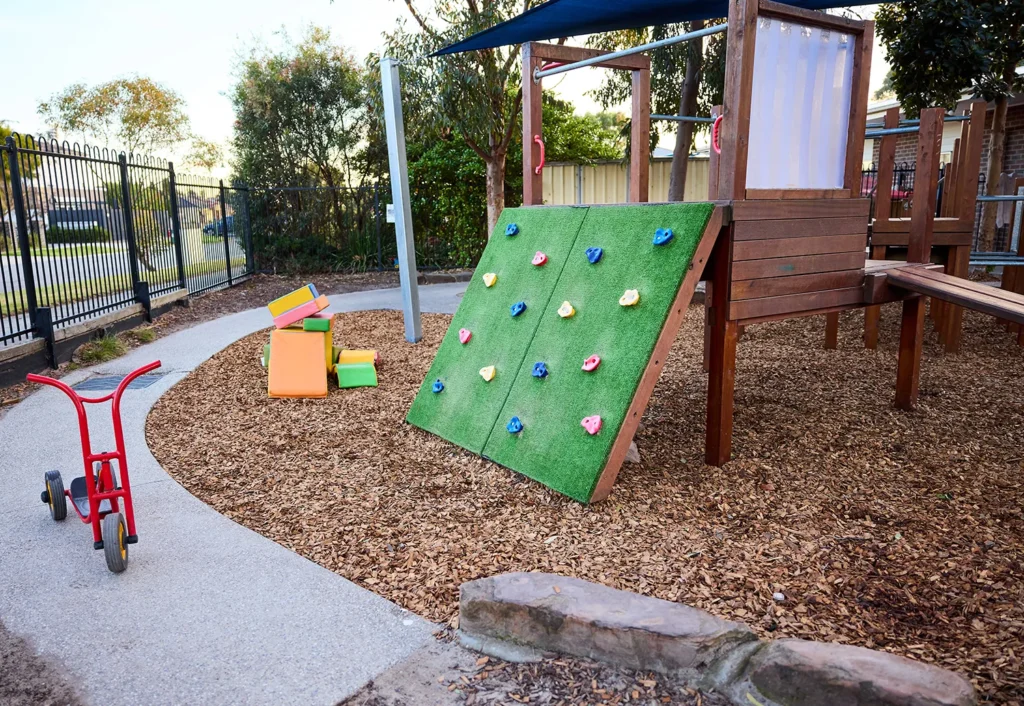 A preschool playground with a wooden climbing structure, a slide, a balancing beam, colorful blocks, and a red tricycle on a gravel path is surrounded by a metal fence and trees, offering an ideal early learning environment.