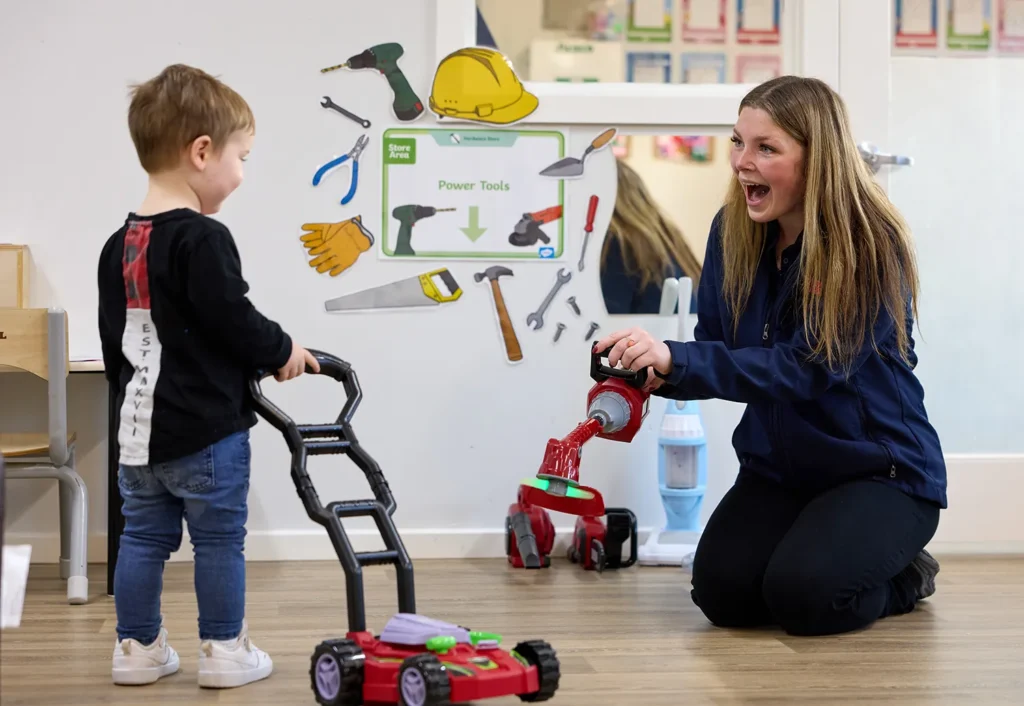 A woman kneels and interacts with a child holding a toy lawn mower in a room adorned with an "Early Learning" educational poster. Toys and a vacuum cleaner are visible, capturing the essence of engaging childcare.