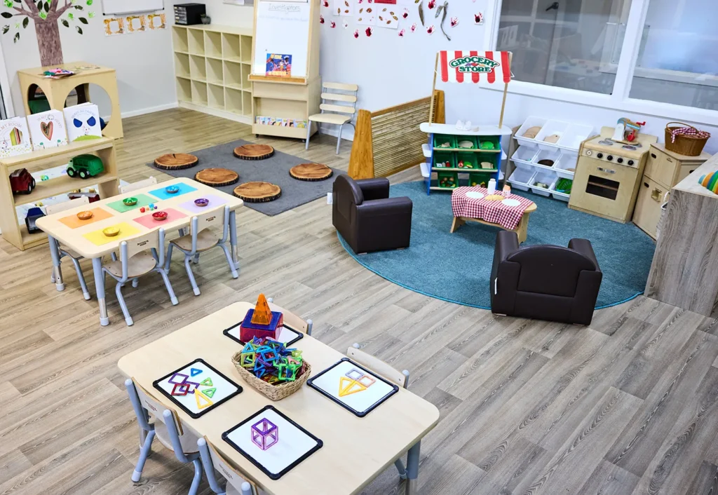 A brightly lit classroom designed for early learning, featuring tables, chairs, educational toys, and learning stations. The room includes a tree stump sitting area for storytime, an art station with paints to inspire creativity, and a play kitchen perfect for imaginative play in kindergarten.