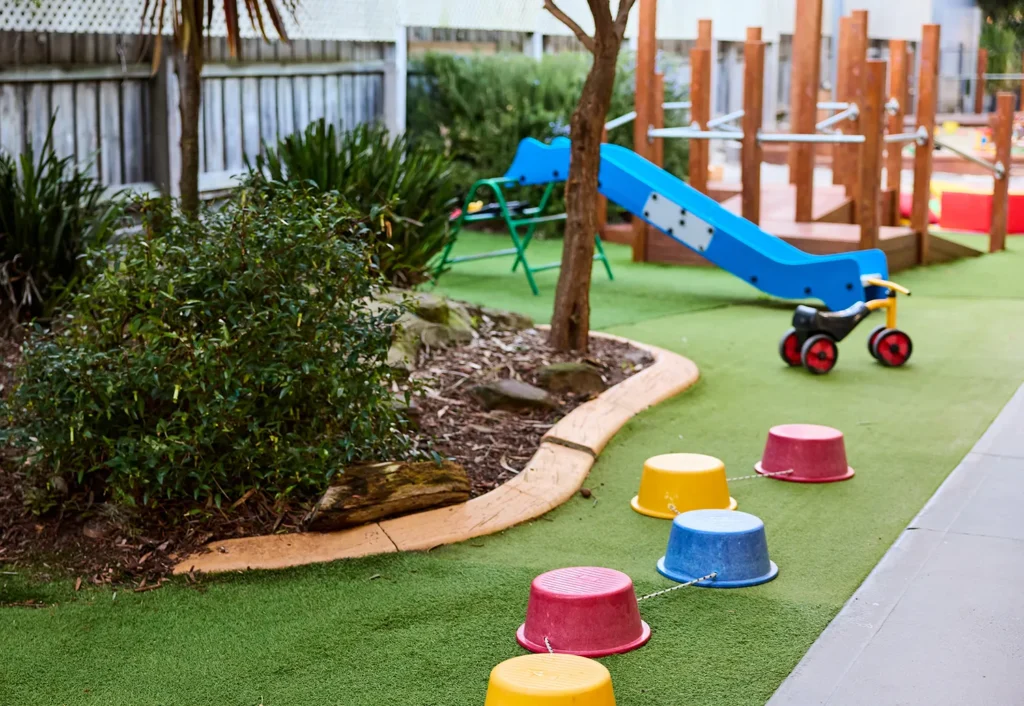 Preschool outdoor play area with a blue slide, climbing equipment, a toy tricycle, multi-colored stepping stones, and greenery.