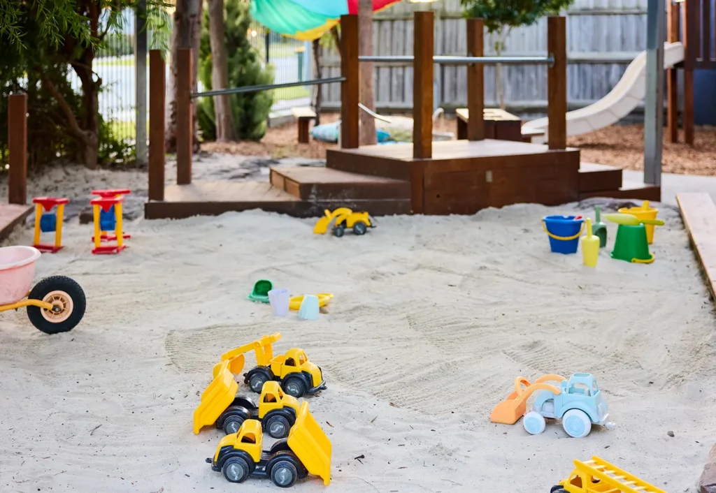 A playground sandbox filled with various toy trucks, buckets, and shovels, perfect for early learning adventures. In the background, a wooden play structure and slides provide additional fun for the preschool day care environment.