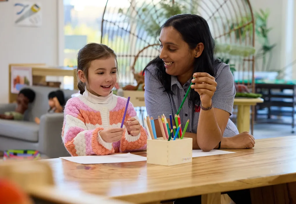 A young girl and an adult are sitting at a table drawing with colored pencils. The adult, embodying the nurturing essence of childcare, is smiling and assisting the child.