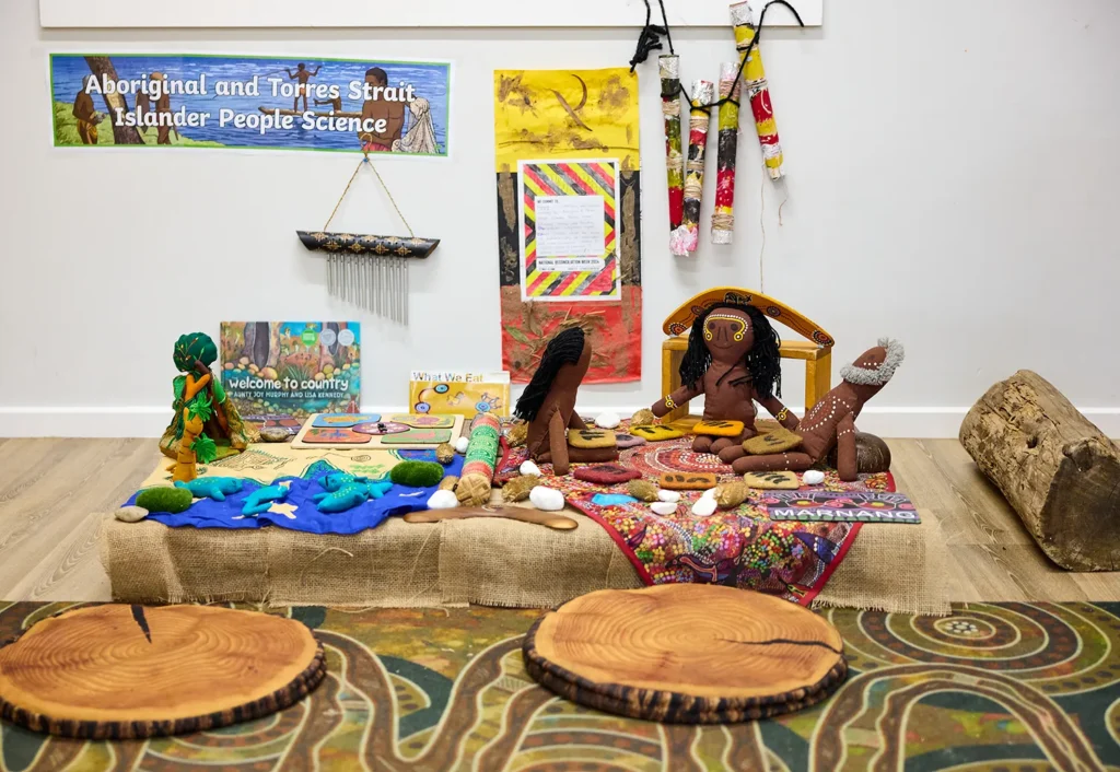 A display at the preschool showcases Aboriginal and Torres Strait Islander cultural artifacts, with books, woven items, dolls, and colorful fabrics arranged on a table. A poster in the background reads, "Aboriginal and Torres Strait Islander People Science.