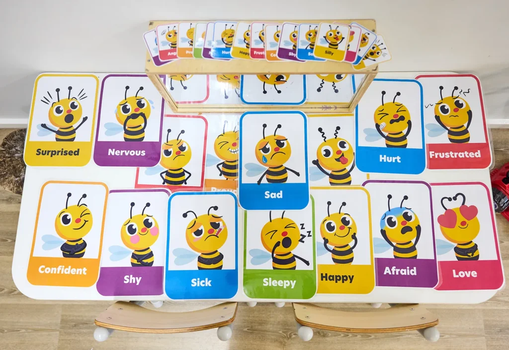 Educational cards with bee illustrations displaying various emotions, including Surprised, Nervous, Confident, Sad, Happy, and more, are arranged on a table—perfect for day care and early learning environments.