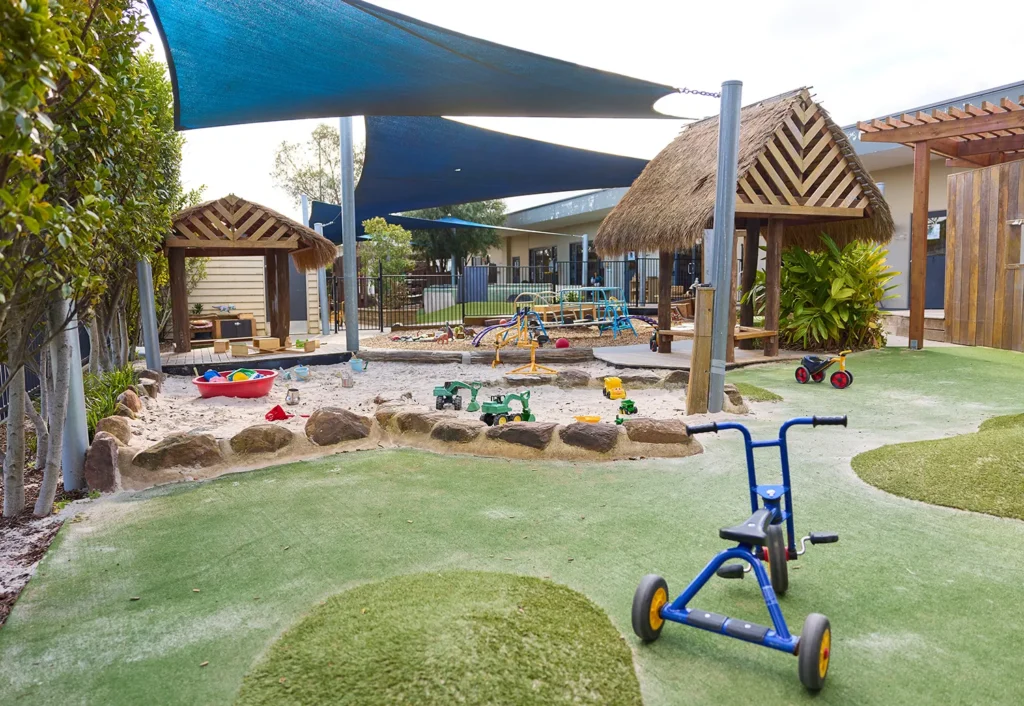 Outdoor playground featuring a sandpit, play structures with thatched roofs, a tricycle, and various toys on an artificial grass surface under sun shade sails, ideal for early learning at day care or kindergarten.