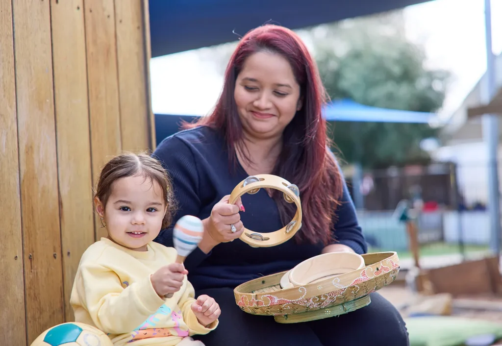 A woman and a young girl sit outdoors at a day care, playing musical instruments including a tambourine and a maraca. The girl is seated, holding the maraca, while the woman sits beside her, smiling and looking at the instruments.