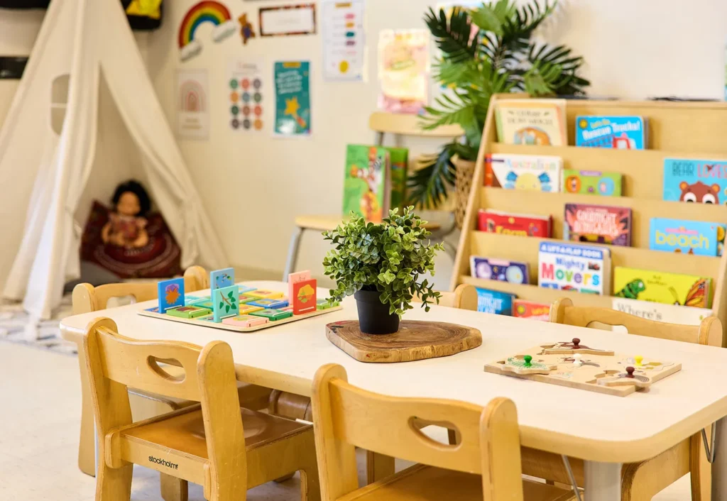 A classroom with a small table and chairs, children's books on a shelf, educational toys, a potted plant on the table, and a play teepee area in the background creates an inviting day care environment.