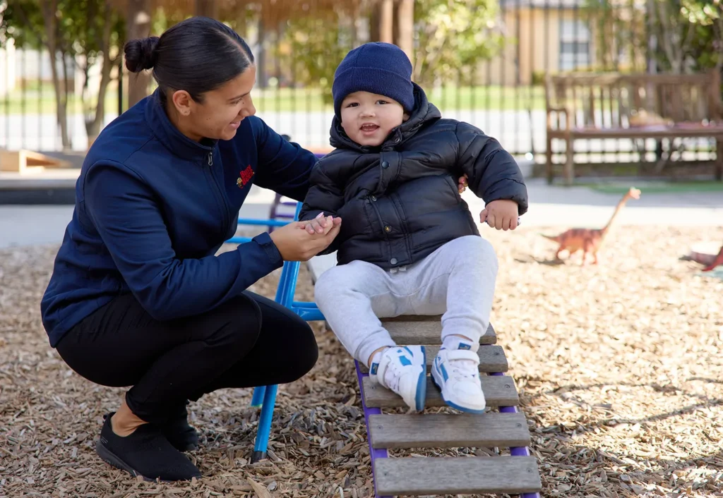 A woman helps a child in a puffy jacket and beanie hat down a small outdoor ramp at the daycare playground.