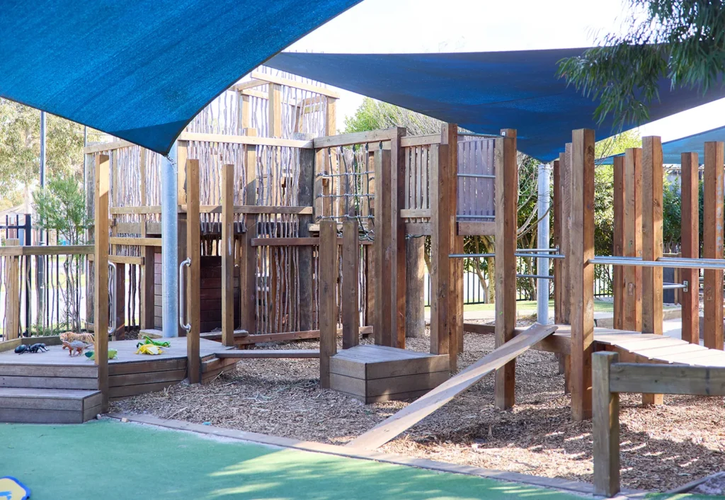 Outdoor playground with wooden climbing structures and a green padded area under blue shade sails. Ideal for kindergarten activities, wood chips cover the ground. Trees and foliage are visible in the background, creating a perfect setting for childcare fun.