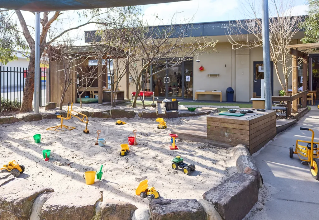 An outdoor playground at the day care features a sandbox filled with toys including trucks, shovels, and buckets. Situated in front of the building, the area is surrounded by trees and benches, offering a perfect setting for early learning and play.