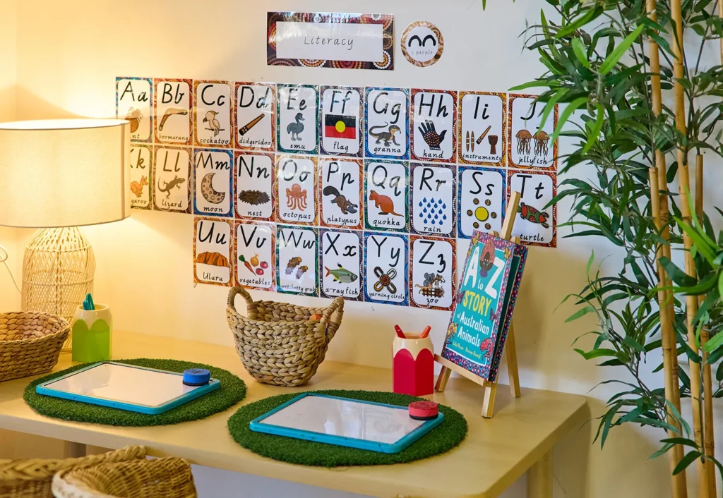 A children's study desk with two whiteboards, markers, and woven baskets. The wall features a colorful alphabet chart with animal illustrations, ideal for early learning at home or in kindergarten. A small plant on the right adds a touch of nature.