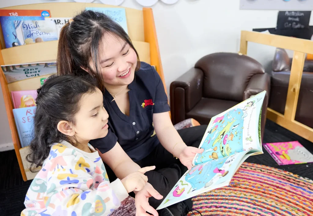 A Busy Bees educator and a child are seated on a colorful rug in a children's reading area, looking at a picture book together.