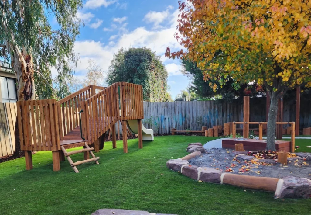 A wooden play fort with a slide in the outdoor play area of Epping daycare, complemented by an Autumn tree.