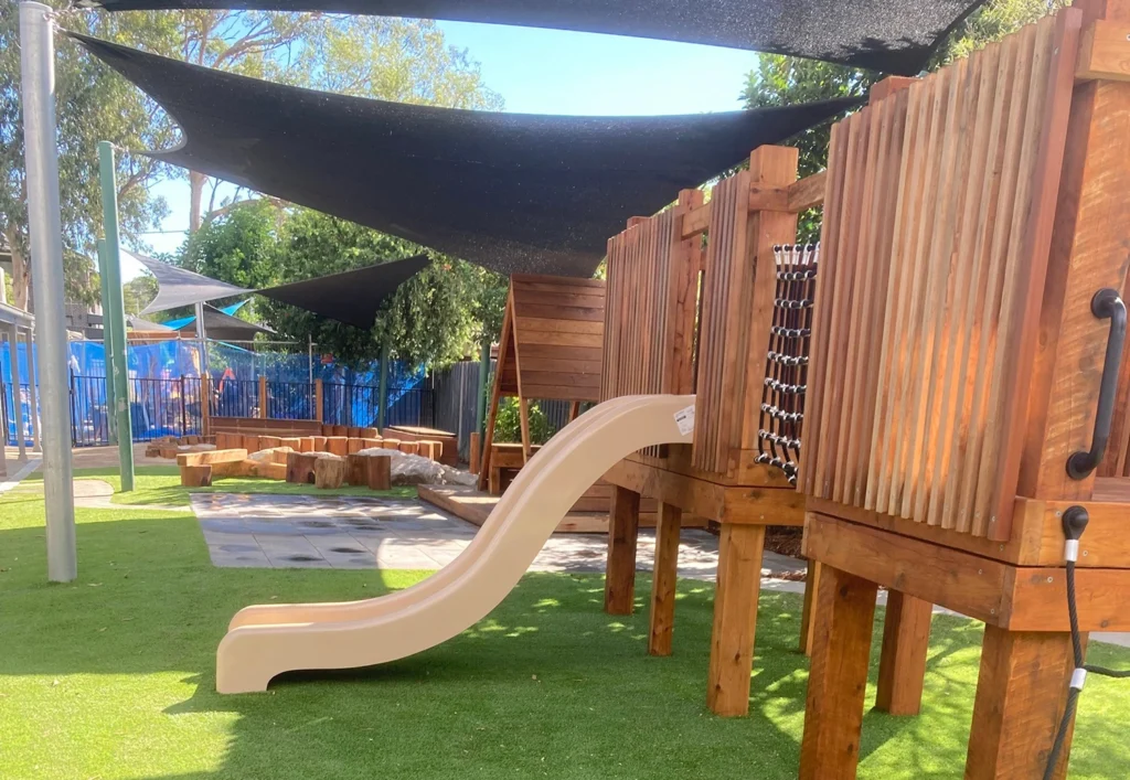 Outdoor playground with wooden structures, a slide, rope net, stepping logs, and artificial grass under shaded cover. Trees and fencing in the background.