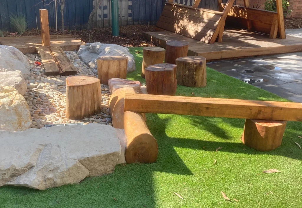 Outdoor playground area with wooden stumps and logs arranged on artificial grass and pebbles, adjacent to a wooden play structure.