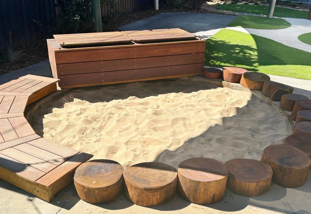 A rectangular sandbox with a wooden cover and wooden log seats around it is located in a playground with a synthetic grass area in the background.