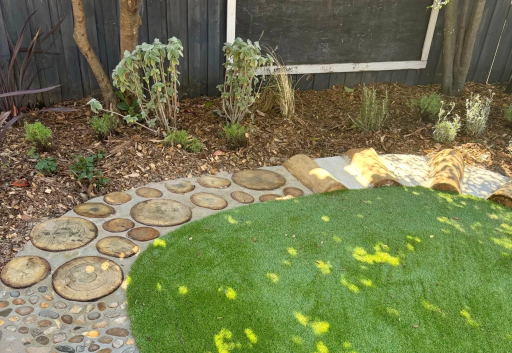 A backyard garden features a path made of circular wooden slices and small stones next to an area of artificial grass, with plants and shrubs in a mulched bed along the fence.