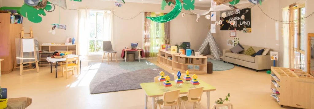 A brightly lit classroom with various seating areas, artwork on walls, bookshelves, and educational toys. There is a play tent, rug, and green decorations hanging from the ceiling.