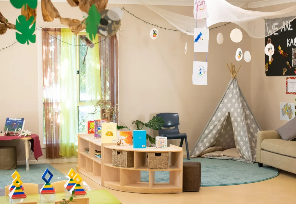 A cozy classroom corner with a play teepee, books on display, a couch, a chair, and various educational toys arranged on a low shelf unit. Wall decorations include hanging artwork and a colorful play mat.