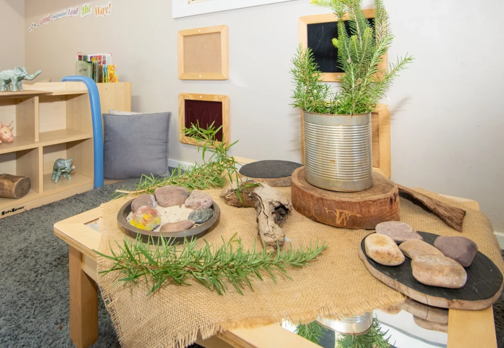 A small wooden table with natural decorations including rocks, wooden slices, and potted plants in a child’s play area. Shelves with toys and a cushion are visible in the background.