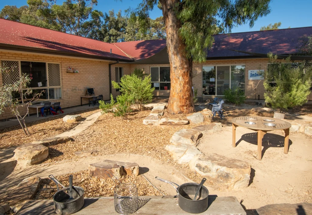 A courtyard features a sand and rock play area surrounded by a beige brick building. Cookware lies on a table in the foreground. Trees and bushes are scattered around the space.