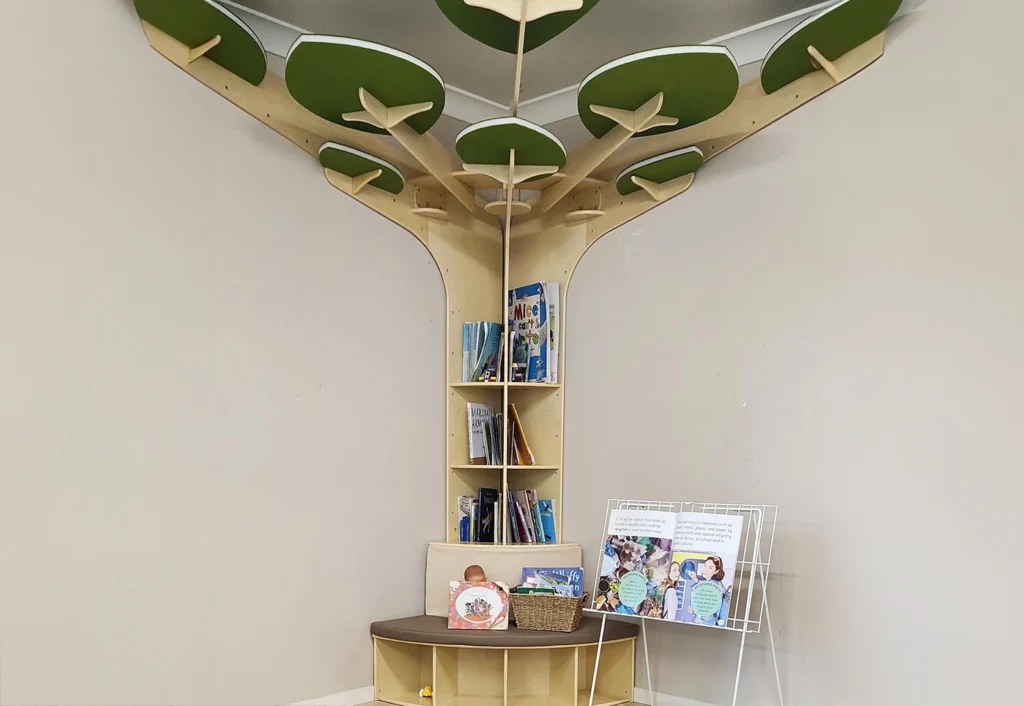 A built-in bookshelf shaped like a tree in the corner of a room, holding books and a basket. A display easel with posters is positioned next to it.