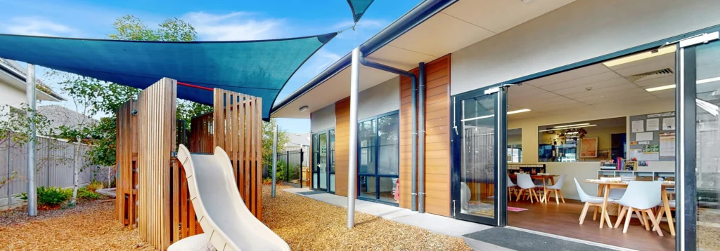 Outdoor view of a modern childcare facility with a play area featuring a slide and shade sail, adjacent to an indoor space with tables and chairs.