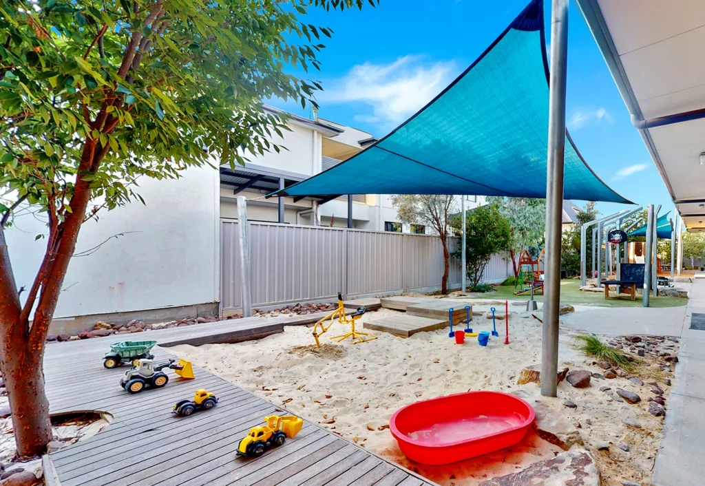 Outdoor playground with a sandy area featuring various toy trucks, a seesaw, and a red bowl. A shade cloth is overhead, and there are adjacent buildings and trees.