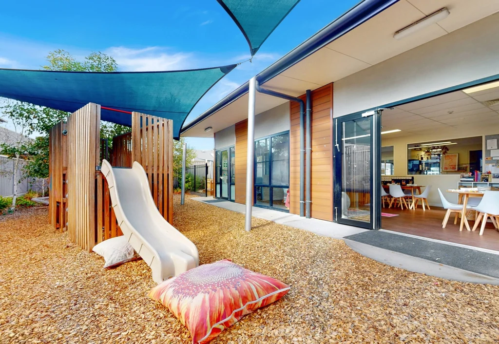 Outdoor play area with a slide and shade sails at a daycare center. Inside, there are tables, chairs, and children's art displayed on the walls.