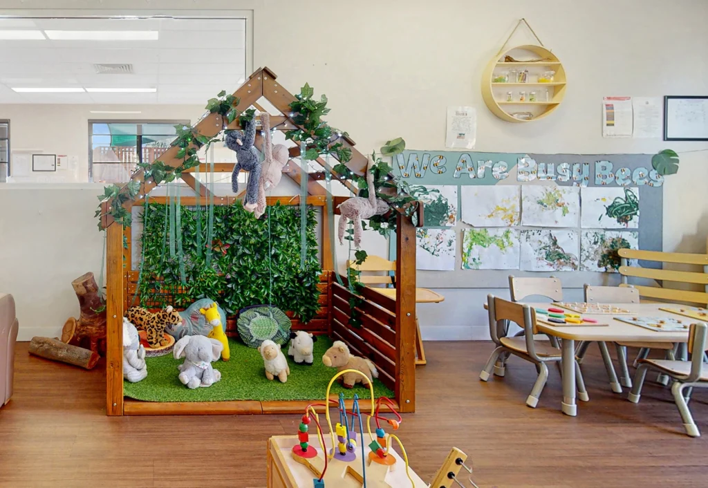 A preschool classroom featuring a play area with animal plush toys, a wooden playhouse, a table with art supplies, and educational posters on the wall.