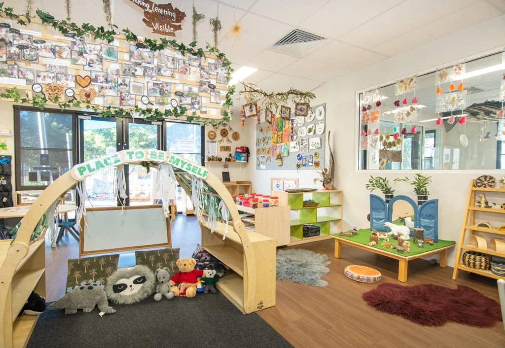 A brightly decorated classroom features children's artwork, educational posters, a reading nook, and various toys and learning stations, with a sign that reads, "PLACE TO BE MYSELF.