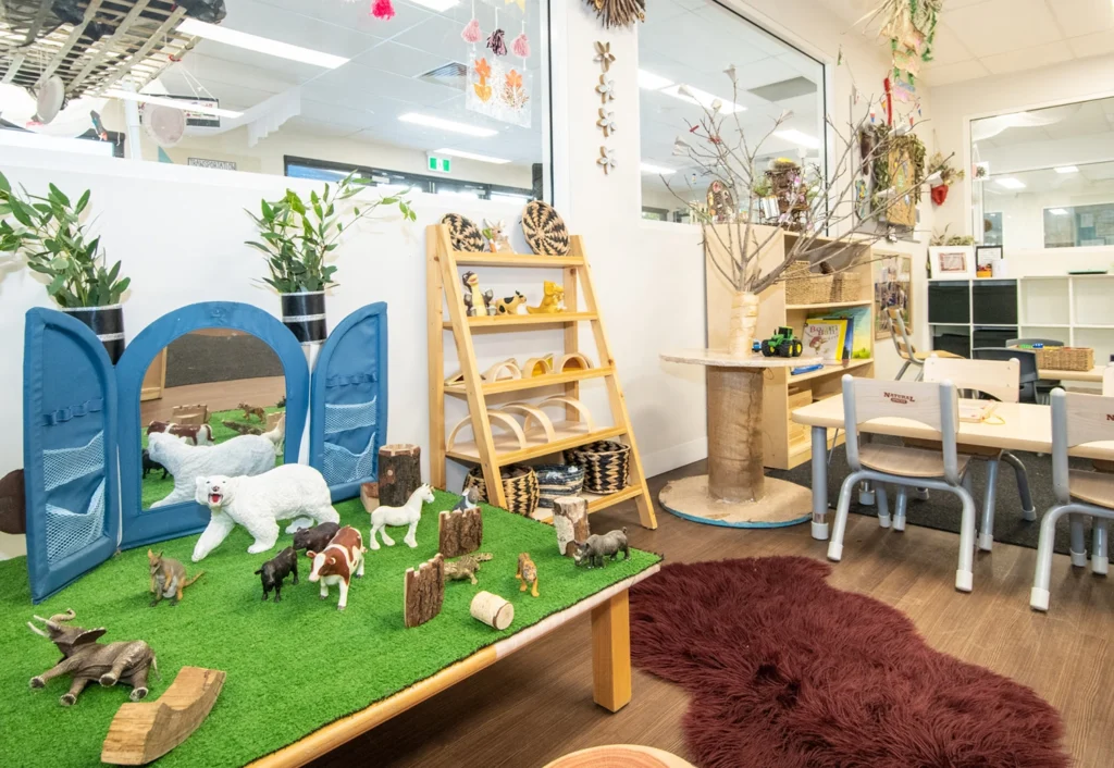 Classroom with animal figures on a play table, shelves with toys, chairs around a table, and wall decorations featuring natural elements.