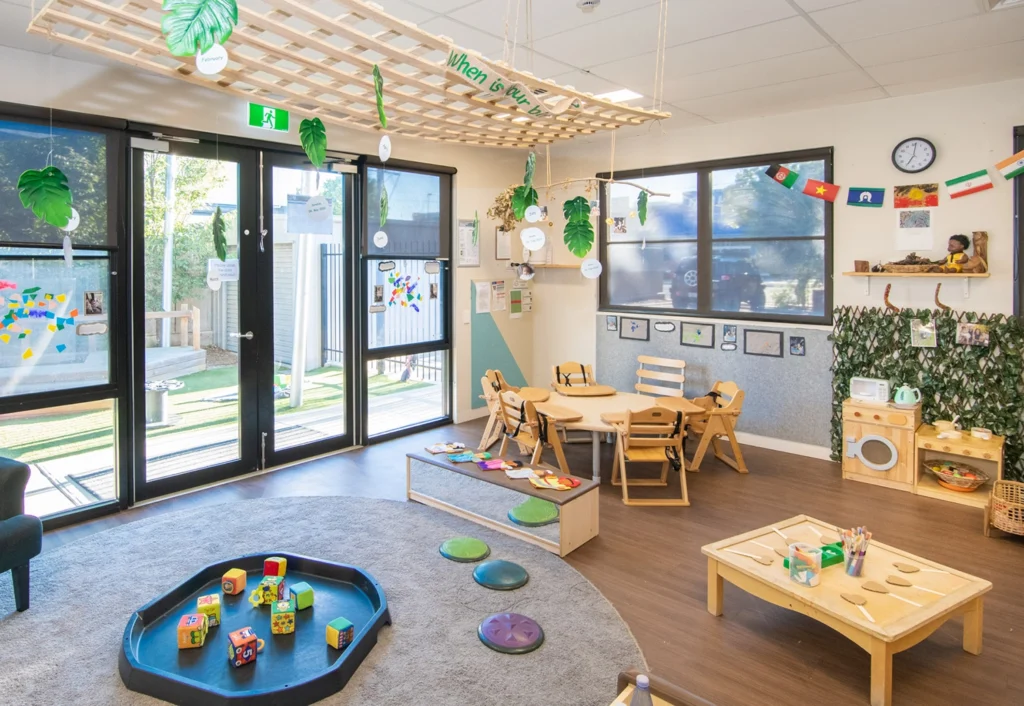 A brightly lit classroom features small tables and chairs, a play area with toys, educational materials on the walls, and large windows letting in natural light.