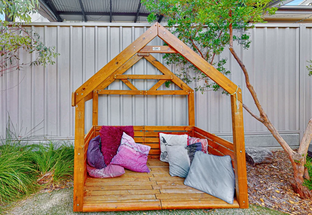 A wooden outdoor playhouse with an open frame design, housing several colorful pillows and cushions, is situated in a garden area.