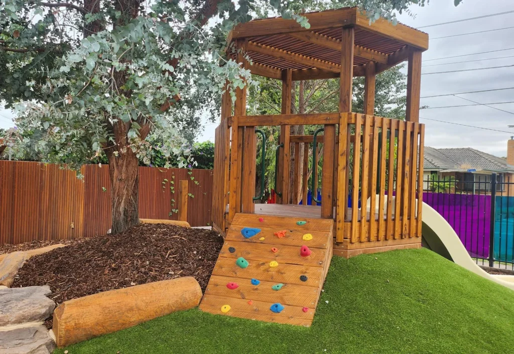 A preschool playground structure featuring a wooden tower with a climbing wall and a slide, set on a grassy mound near a tree and surrounded by a fence.