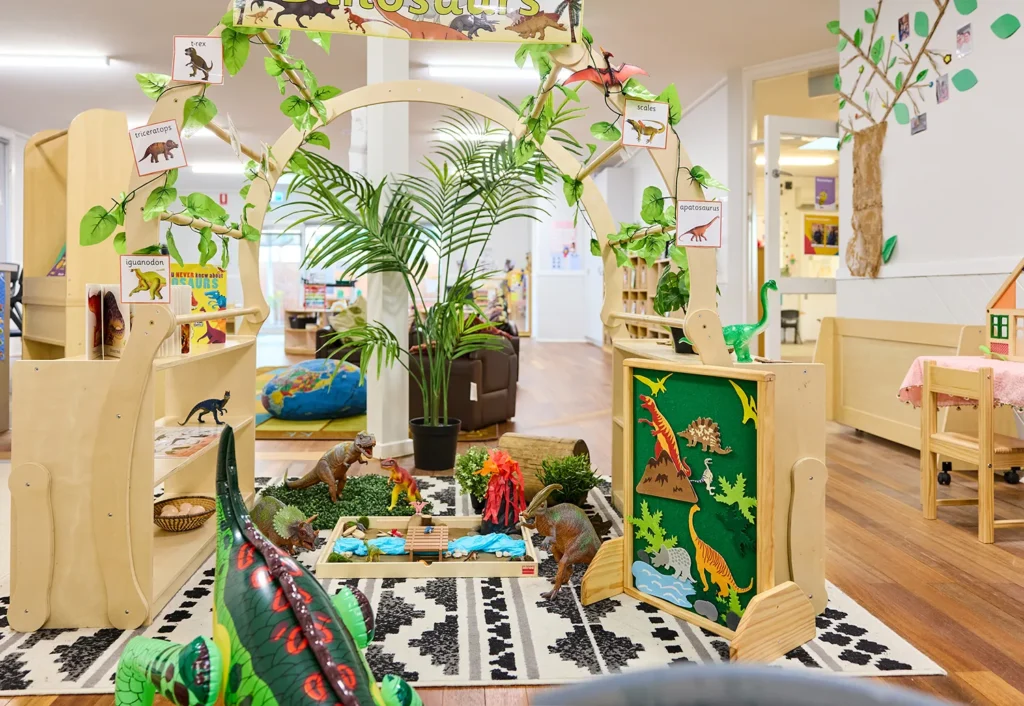 Classroom dinosaur-themed play area with wooden arches, toy dinosaurs, a felt board with dinosaur figures, and a small volcano model, set on a patterned rug with surrounding educational decor designed for preschool early learning.