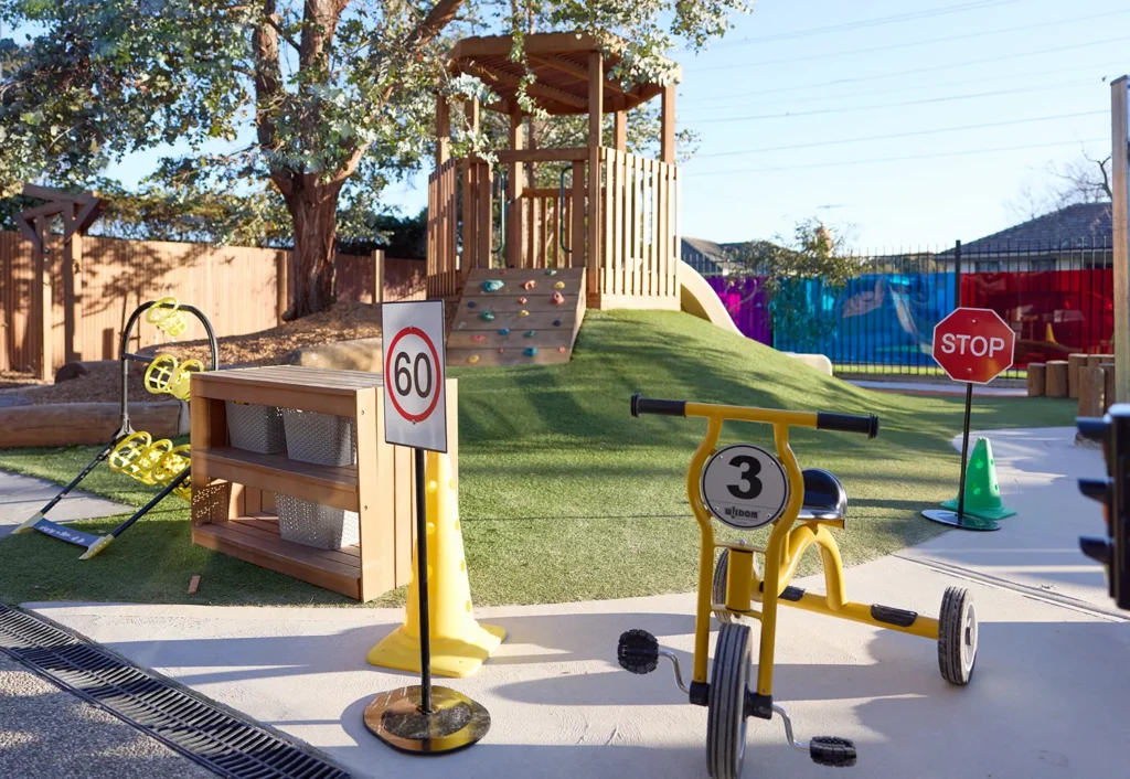 In the kindergarten's outdoor play area, children can enjoy a climbing structure, toy traffic signs, a yellow tricycle, and a pretend gas station. Surrounded by trees, fences, and colorful walls, it's an ideal space promoting early learning and fun.