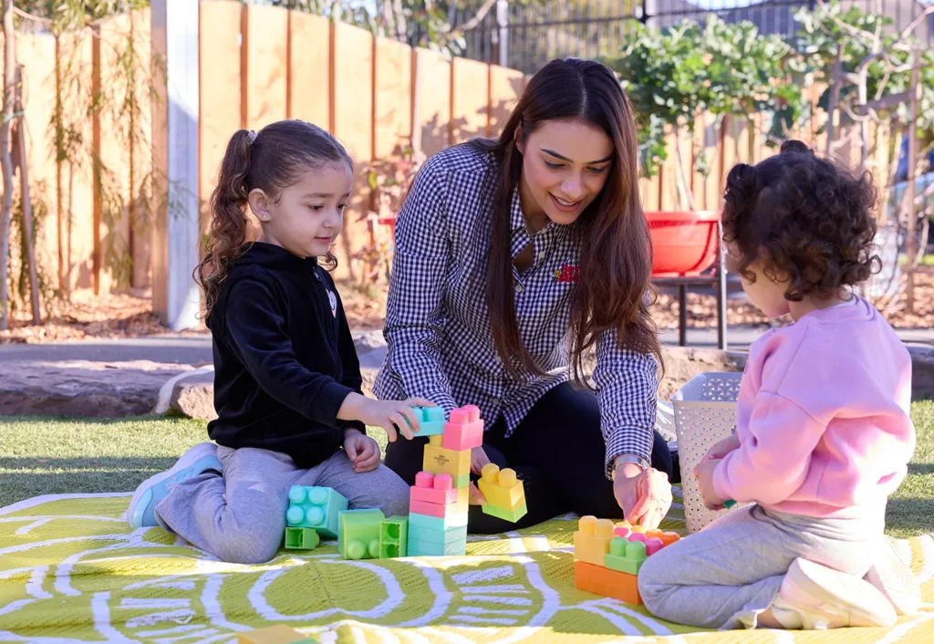 A woman and two children are sitting on a yellow blanket outside, building structures with large colorful plastic blocks in a lively kindergarten setting.