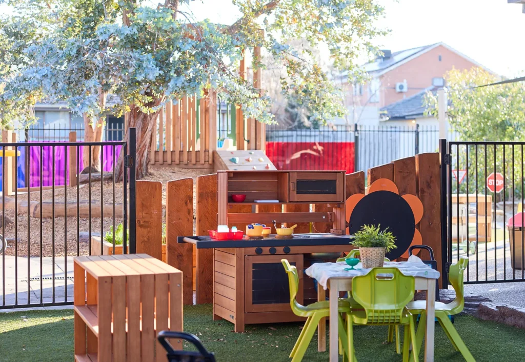 An outdoor playground at the day care features a wooden play kitchen set, green chairs, and a table. A tree, fence, and buildings are in the background. Colorful panels add a vibrant touch to this delightful setting where children thrive.