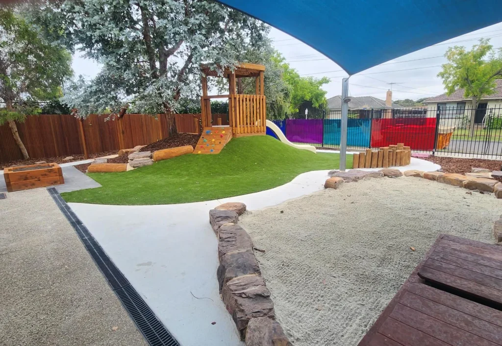 The outdoor playground, ideal for a kindergarten or early learning center, features artificial turf, a wooden play structure, a sandpit with stone borders, and colorful fencing. A large blue shade cover provides protection from the sun.