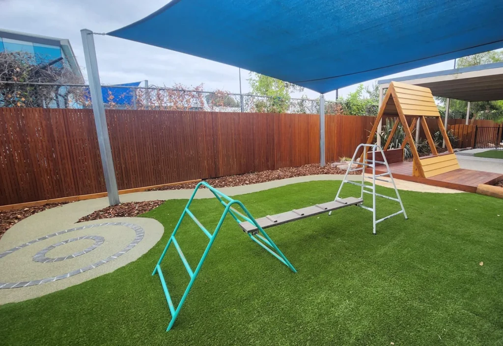 An outdoor playground at the preschool features a climbing structure on artificial grass, shaded by a blue canopy.